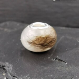 Puede incluir: Una cuenta redonda y blanca con un anillo de metal plateado en la parte superior. La cuenta tiene una secci&oacute;n de material marr&oacute;n similar al cabello. La cuenta est&aacute; sobre una superficie oscura y texturizada.