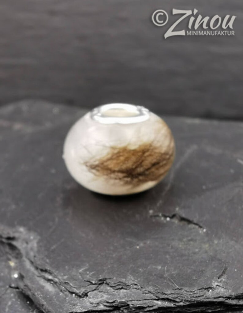 Puede incluir: Una cuenta redonda y blanca con un anillo de metal plateado en la parte superior. La cuenta tiene una secci&oacute;n de material marr&oacute;n similar al cabello. La cuenta est&aacute; sobre una superficie oscura y texturizada.