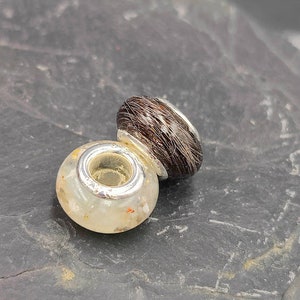 May include: Two decorative beads. One is a clear, round bead with flecks of color and a silver-colored metal ring. The other is a dark brown, oval-shaped bead with a silver-colored metal ring.
