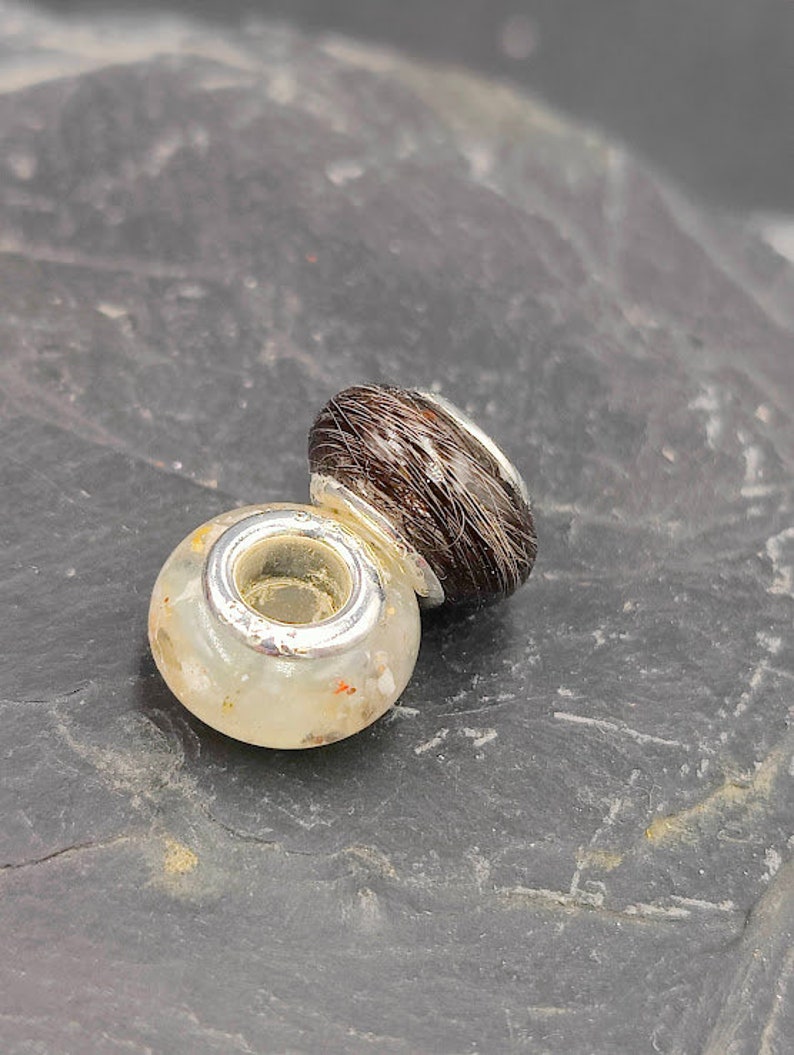 May include: Two decorative beads. One is a clear, round bead with flecks of color and a silver-colored metal ring. The other is a dark brown, oval-shaped bead with a silver-colored metal ring.