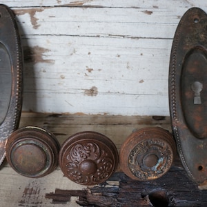 Może przedstawiać: Asortyment zabytkowych okuć do drzwi, w tym dwie owalne płytki tylne i trzy gałki. Okucia wykonane są ze starego metalu z rdzawobrązowym wykończeniem, wykazującym oznaki zużycia. Gałki mają różne dekoracyjne wzory.