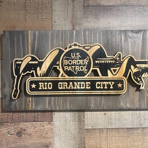Può includere: Un cartello in legno con un design nero e marrone. Il cartello presenta un'immagine stilizzata di un serpente, un fucile e le parole "U.S. Border Patrol" e "Rio Grande City".