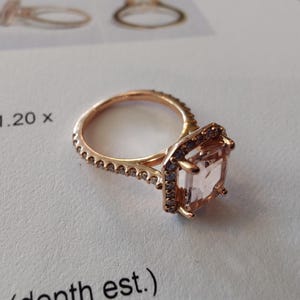 May include: A rose gold engagement ring with a square-shaped, light pink gemstone surrounded by a halo of smaller diamonds. The band is also set with smaller diamonds.