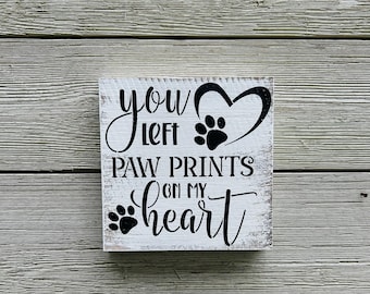 May include: A square, white wooden sign with black text and paw prints. The sign reads "You left paw prints on my heart" in a decorative font. A heart shape with a paw print is above the words.