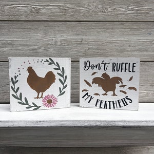 May include: Two wooden signs with a farmhouse theme. The first sign features a brown chicken silhouette with a pink flower and green leaves. The second sign says "Don't ruffle my feathers" with a brown rooster silhouette and feathers.