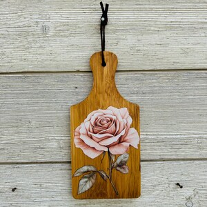 May include: Wooden cutting board with a handle and a detailed illustration of a pink rose. The rose has multiple petals and green leaves. A dark brown leather cord is attached to the handle for hanging.