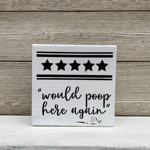May include: A white wooden sign with black text that reads "would poop here again" and five black stars above the text.