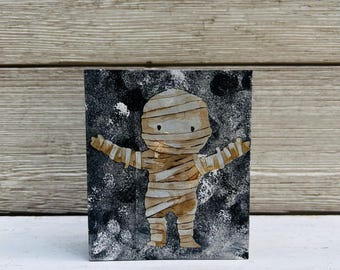 May include: A small wooden block with a black background and a white mummy illustration. The mummy is wearing bandages and has a simple face with two eyes.