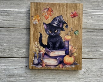 May include: Wooden sign featuring a black cat wearing a witch hat, sitting atop a stack of books. The artwork includes pumpkins, autumn leaves, and small bottles, creating a Halloween or fall theme. The background has a wood grain texture.