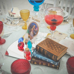 May include: A table setting with a red wine glass, a gold goblet, and a blue glass vase. There are also several books stacked on top of each other, a small mirror, and a card with the word "Dalaran" on it.
