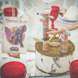 May include: Two wooden totem poles with painted designs stand on a birch log base covered in moss. The totem poles have wings and are painted in shades of pink, blue, and green. The text "THUNDER BLUFF" is visible on a piece of paper in the background.