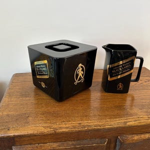 May include: A black square ice bucket and a matching black pitcher, both featuring the gold Johnnie Walker Black Label logo and the iconic striding person figure. The ice bucket includes a lid, and both items display the 12-year age statement.
