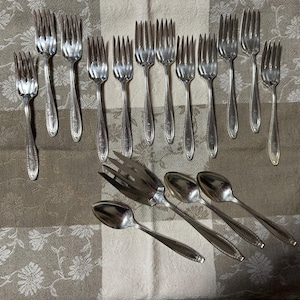 May include: A set of silverware, including forks and spoons, laid out on a white and brown patterned cloth. The silverware is made of silver and has a polished finish.
