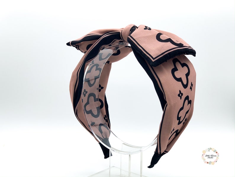 May include: A pink and black patterned headband with a large bow. The headband is made of fabric and has a black elastic band.