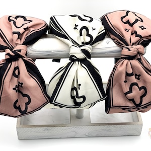 May include: Three fabric headbands with a black and white geometric pattern and a pink and black geometric pattern. The headbands are tied in a bow and displayed on a white stand.
