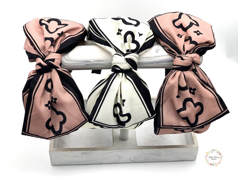 May include: Three fabric headbands with a black and white geometric pattern and a pink and black geometric pattern. The headbands are tied in a bow and displayed on a white stand.