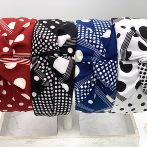 May include: A collection of knotted headbands in various colors and patterns. The headbands feature a mix of polka dots and geometric designs in red, black, white, and blue. The headbands are displayed on a white stand.
