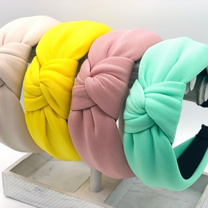 Può includere: Quattro fasce per capelli annodate in diversi colori: beige, giallo, rosa e verde menta. Le fasce sono esposte su un supporto bianco.