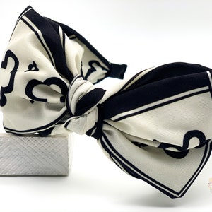 May include: A white and black patterned headband with a large bow. The bow is tied in a knot and the headband is made of a soft fabric.