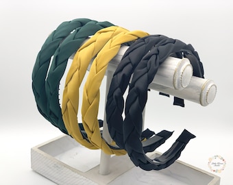 May include: Four braided headbands in different colors: dark green, mustard yellow, black, and a lighter shade of yellow. The headbands are displayed on a white stand.
