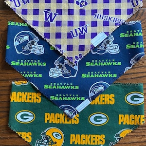 May include: Three dog bandanas with different team logos. The top bandana is purple and yellow with the text "UW Huskies" and a paw print. The middle bandana is blue with the text "Seattle Seahawks" and a football helmet. The bottom bandana is green with the text "Packers" and a football helmet.