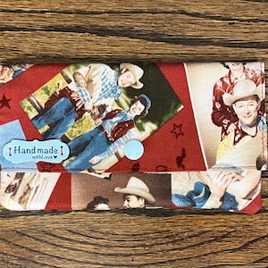 May include: A red fabric wallet with a collage of vintage photos of cowboys and cowgirls. The wallet has a blue button closure and a small tag that says "Handmade with love".