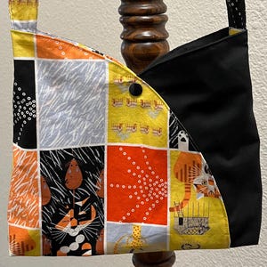 May include: A patchwork shoulder bag with a black, orange, yellow, and gray cat-themed design. The bag features a black panel and a button closure. The strap is yellow with black polka dots.