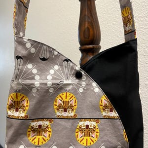 May include: A gray and black fabric tote bag with a cat pattern. The bag has a long strap and a black flap with a button closure.
