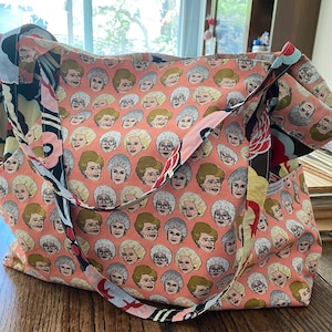 May include: A pink tote bag with a repeating pattern of a woman's face. The bag has black straps and a black and white floral pattern on the side.