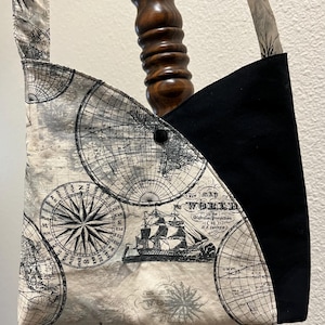 May include: A black and white fabric tote bag with a map of the world print. The bag has a black strap and a black flap with a compass design. The bag has a button closure.
