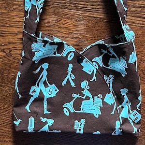 May include: A brown fabric tote bag with blue silhouettes of women and scooters. The bag has a long strap and a button closure.