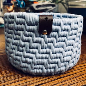 May include: A light blue, hand-knitted basket with a woven texture. A brown leather strap with a silver snap closure is attached to the side. The basket is round and sits on a wooden surface.
