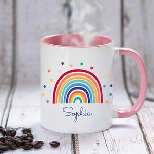 May include: White ceramic mug with a pink rim and handle. The mug features a colorful rainbow design with a scattering of colorful dots. The name "Sophia" is written in blue cursive below the rainbow.