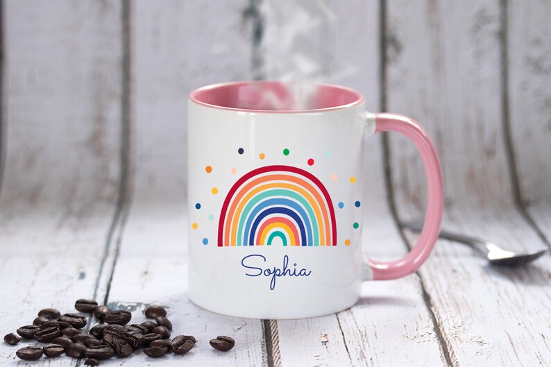 May include: White ceramic mug with a pink rim and handle. The mug features a colorful rainbow design with a scattering of colorful dots. The name "Sophia" is written in blue cursive below the rainbow.