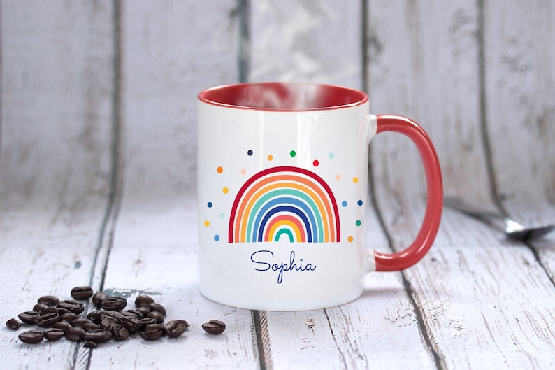 May include: White ceramic mug with a red rim and handle. The mug features a colorful rainbow design with a scattering of polka dots and the name "Sophia" printed below.