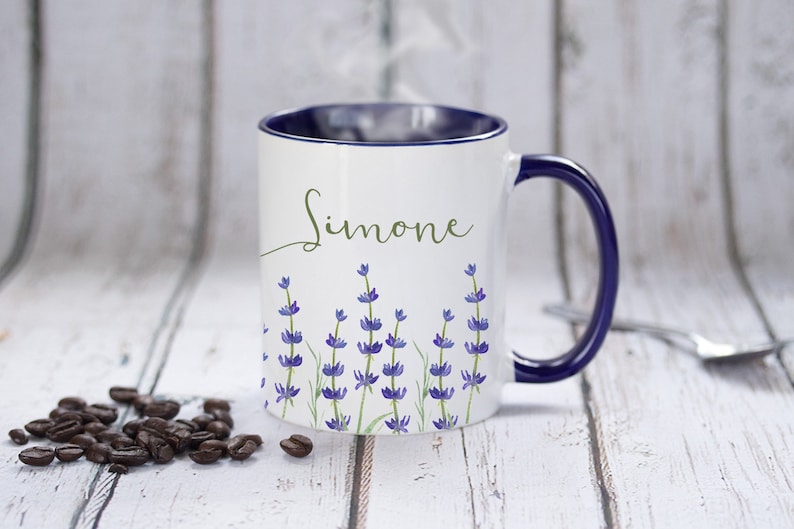 May include: White ceramic mug with a blue rim and handle. The mug has a lavender floral design and the name "Simone" written in cursive on the front.