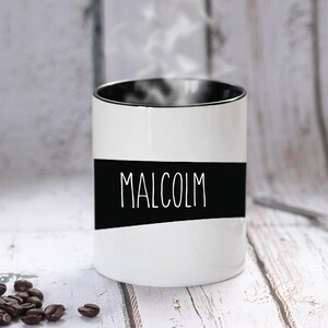 May include: A white ceramic mug with the name "MALCOLM" in white lettering on a black band. The mug has a black interior and is emitting steam. Coffee beans are scattered near the mug.