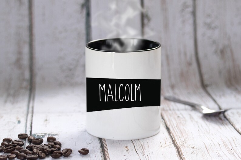 May include: A white ceramic mug with the name "MALCOLM" in white lettering on a black band. The mug has a black interior and is emitting steam. Coffee beans are scattered near the mug.
