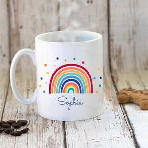 May include: White ceramic mug with a rainbow design and colorful polka dots. The name "Sophia" is printed below the rainbow.