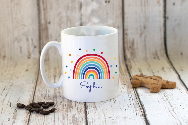 May include: White ceramic mug with a rainbow design and colorful polka dots. The name "Sophia" is printed below the rainbow.