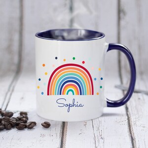 May include: White ceramic mug with a blue rim and handle. The mug features a colorful rainbow design with a scattering of multi-colored dots and the name "Sophia" printed below the rainbow.
