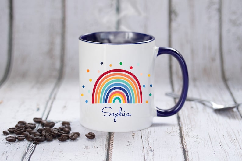 May include: White ceramic mug with a blue rim and handle. The mug features a colorful rainbow design with a scattering of multi-colored dots and the name "Sophia" printed below the rainbow.