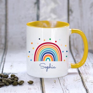May include: A white ceramic mug with a yellow rim and handle. The mug features a colorful rainbow design with a scattering of dots and the name "Sophia" printed below.
