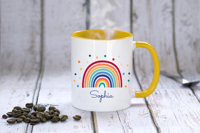 May include: A white ceramic mug with a yellow rim and handle. The mug features a colorful rainbow design with a scattering of dots and the name "Sophia" printed below.
