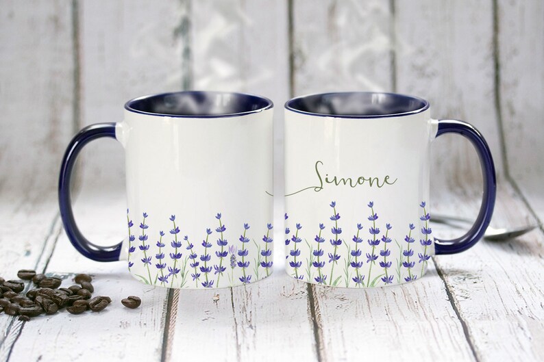 May include: Two white ceramic mugs with blue rims and handles. One mug has the name "Simone" written in cursive on the front. Both mugs feature a lavender floral design.