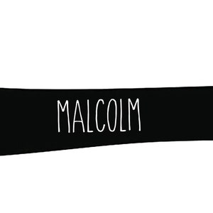 May include: A black silhouette of a dachshund dog with the name "MALCOLM" in white, centered on the dog's body. The dog is in a standing position, with its legs visible and tail slightly curved. The background is plain white.