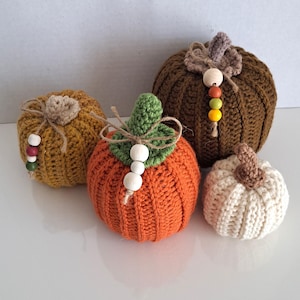 May include: Four crocheted pumpkins in different sizes and colours. The largest pumpkin is brown, the next largest is orange, the next is yellow, and the smallest is white. Each pumpkin has a stem and wooden beads.