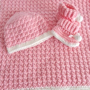 Pode incluir: Um cobertor de bebé de crochê rosa com um chapéu e sapatinhos combinando. O cobertor tem uma borda branca e o chapéu e os sapatinhos têm uma guarnição branca.
