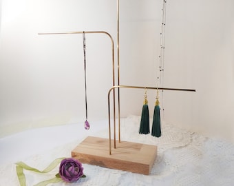 Könnte beinhalten: Ein goldfarbener Schmuckständer aus Metall mit einer Holzbasis. Der Ständer hat drei Arme zum Aufhängen von Halsketten und Ohrringen. Ein grüner Quastenohrring und eine violette Anhängerhalskette sind auf dem Ständer ausgestellt.