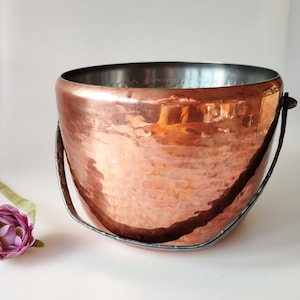May include: A hammered copper ice bucket with a black metal handle. The bucket has a shiny, metallic finish.